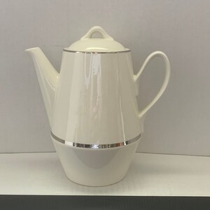 May include: A white ceramic coffee pot with a silver trim around the top and bottom. The pot has a rounded body and a spout on one side.