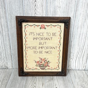 May include: A framed cross-stitch with the text "IT'S NICE TO BE IMPORTANT BUT MORE IMPORTANT TO BE NICE." The embroidery features a floral design in a basket. The frame is dark brown.
