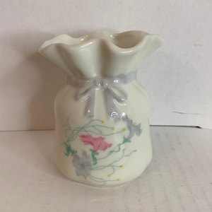 May include: A white ceramic vase shaped like a bag with a ruffled top and a bow. The vase is decorated with a floral design in shades of pink, blue, and green.