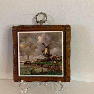 May include: A wooden framed tile with a picture of a windmill in a Dutch landscape. The tile has a metal ring on the top for hanging.