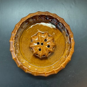 Puede incluir: Un sapo de flores de cerámica marrón con una base circular y una parte superior en forma de estrella con agujeros para sujetar los tallos.