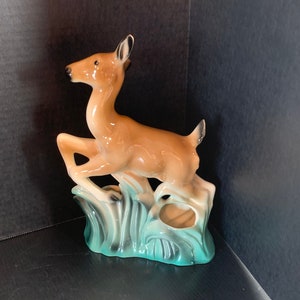 Puede incluir: Una figurita de cerámica de un ciervo con pintura marrón y blanca, de pie sobre una base verde azulada con hierba estilizada.