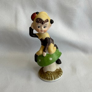 May include: A ceramic figurine of a whimsical elf with brown hair and a yellow and brown outfit, sitting on a green mushroom with white spots.