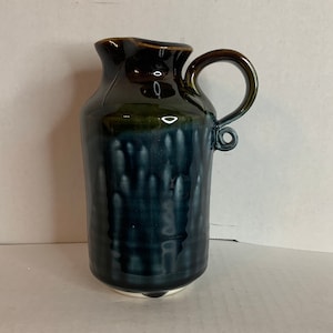 May include: A small, dark green and blue ceramic jug with a white rim and a handle. The jug has a textured surface.
