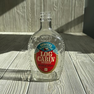 Puede incluir: Una botella de vidrio transparente con una etiqueta roja, blanca y azul que dice "Special Bicentennial Flask Log Cabin Syrup Net 24 FL. OZ. (1 PT 8 OZ)".