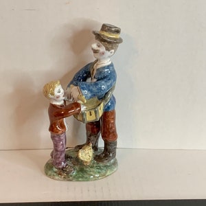 May include: A ceramic figurine of a man and a boy. The man is wearing a blue jacket, brown pants, and a black hat. He is holding a drum. The boy is wearing a red shirt, brown pants, and a yellow hat. The figurine is painted in bright colors.
