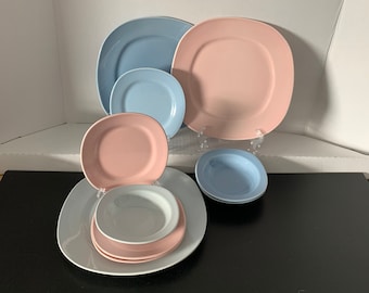 May include: A set of six ceramic plates in various sizes and colors, including pink, blue, and white. The plates are arranged in a pyramid shape on a black surface.