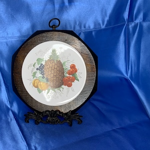May include: A wooden octagonal frame with a white ceramic tile featuring a pineapple, cherries, and grapes. The frame has a black metal stand.