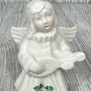 May include: A white porcelain figurine of an angel playing a musical instrument. The angel has a white dress with green shamrocks on the bottom.