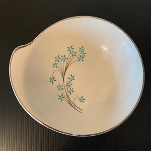 Puede incluir: Un tazón de cerámica blanca con un borde plateado y un diseño floral azul. El tazón tiene una forma única con un borde curvo.