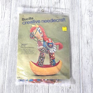 May include: A Bucilla creative needlecraft kit featuring a "Rocky" rocking horse design. The kit includes a pattern and materials to create a 22-inch tall stuffed rocking horse with red, blue, and patterned fabric. The kit is sealed in a clear plastic bag.