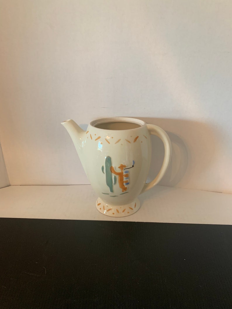 May include: A white ceramic teapot with a brown, green, and blue cactus and person design. The teapot has a handle and a spout.