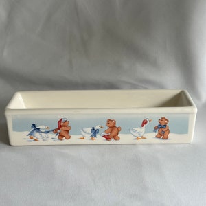 May include: A white ceramic rectangular dish with a blue and white winter scene. The scene features two brown bears wearing red hats and blue bows, and two white geese with blue bows.