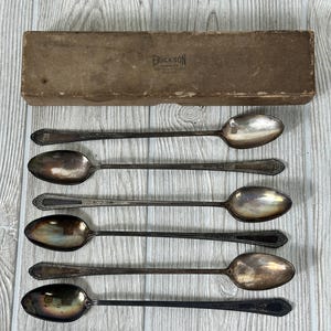 Puede incluir: Juego de seis cucharas de té helado plateadas en una caja marrón con el texto "ERICKSON JEWELER" impreso en la parte superior. Las cucharas tienen un diseño adornado y están en buen estado.