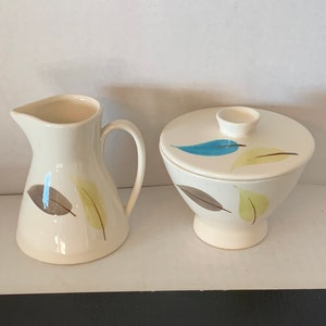 May include: A white ceramic creamer and sugar bowl set with a leaf design in shades of brown, yellow, and blue.