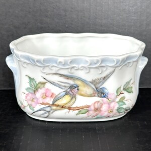May include: A white ceramic planter with blue and white floral designs and two yellow birds with blue accents perched on a branch. The planter has two handles on the sides.