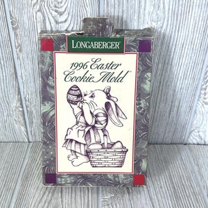 Puede incluir: Una caja morada y verde con un conejo blanco sosteniendo un huevo de Pascua. La caja dice "1996 Easter Cookie Mold" y "Longaberger".