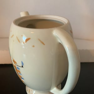 May include: A white ceramic teapot with a brown and blue floral design. The teapot has a handle and a spout. The teapot is sitting on a white surface.