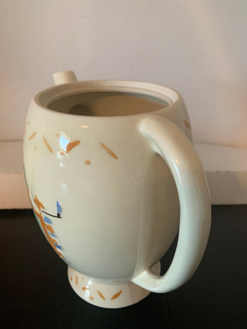 May include: A white ceramic teapot with a brown and blue floral design. The teapot has a handle and a spout. The teapot is sitting on a white surface.