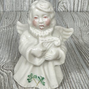 May include: White porcelain figurine of an angel with wings, holding a harp. The angel has rosy cheeks and is wearing a white gown with green shamrocks at the bottom.