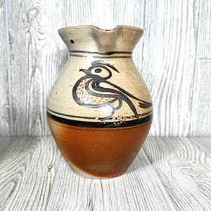 May include: A ceramic pitcher with a rounded body and a flared spout. The upper portion is cream-colored with a hand-painted black line drawing of a bird. A thick black horizontal band separates the cream top from the solid reddish-brown base.