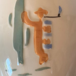 May include: A ceramic figurine of a person holding a stick with a flame on the end. The person is wearing a hat and has a blue and white striped shirt. The figurine is painted in shades of blue, green, and orange.