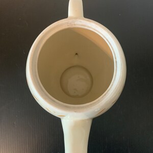 May include: White teapot with a gold pattern. The teapot has a round body and a spout. The interior of the teapot is visible.