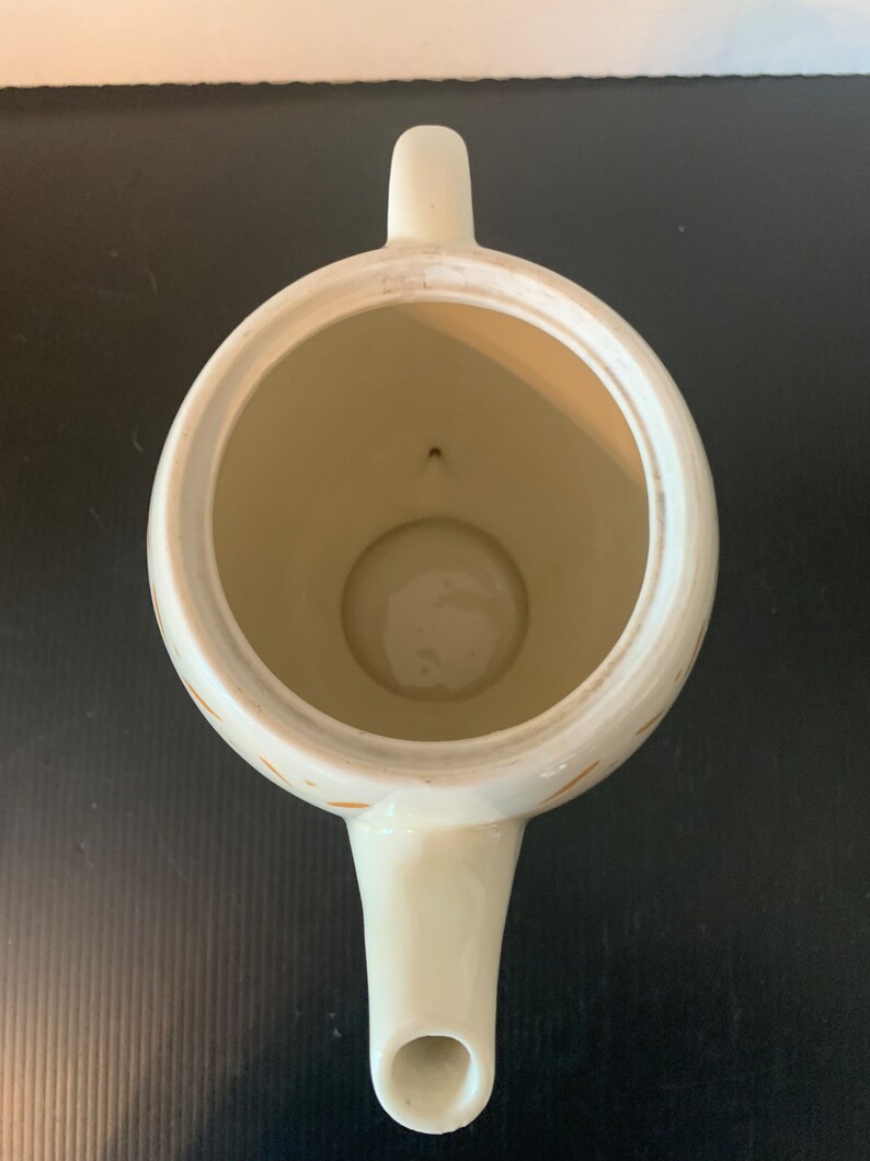 May include: White teapot with a gold pattern. The teapot has a round body and a spout. The interior of the teapot is visible.