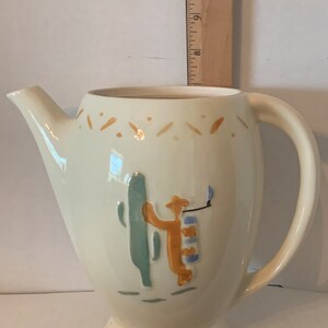 May include: A white ceramic pitcher with a brown, blue, and green design of a person standing next to a cactus. The pitcher has a handle and a spout.