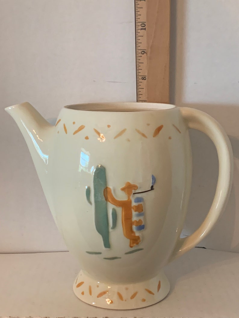 May include: A white ceramic pitcher with a brown, blue, and green design of a person standing next to a cactus. The pitcher has a handle and a spout.