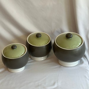 May include: Set of three ceramic canisters with lids. The canisters are brown with a white base and green lids. The lids have a small knob on top.