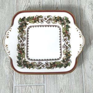 May include: A white rectangular serving platter with a red and gold trim. The platter features a square holly and floral design in the center, with a decorative border and handles.