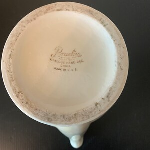 May include: The bottom of a white ceramic pitcher with the words "Porcelaine Since 1921 Vitreous Hand Dec. China Made in U.S.A." printed on the bottom.