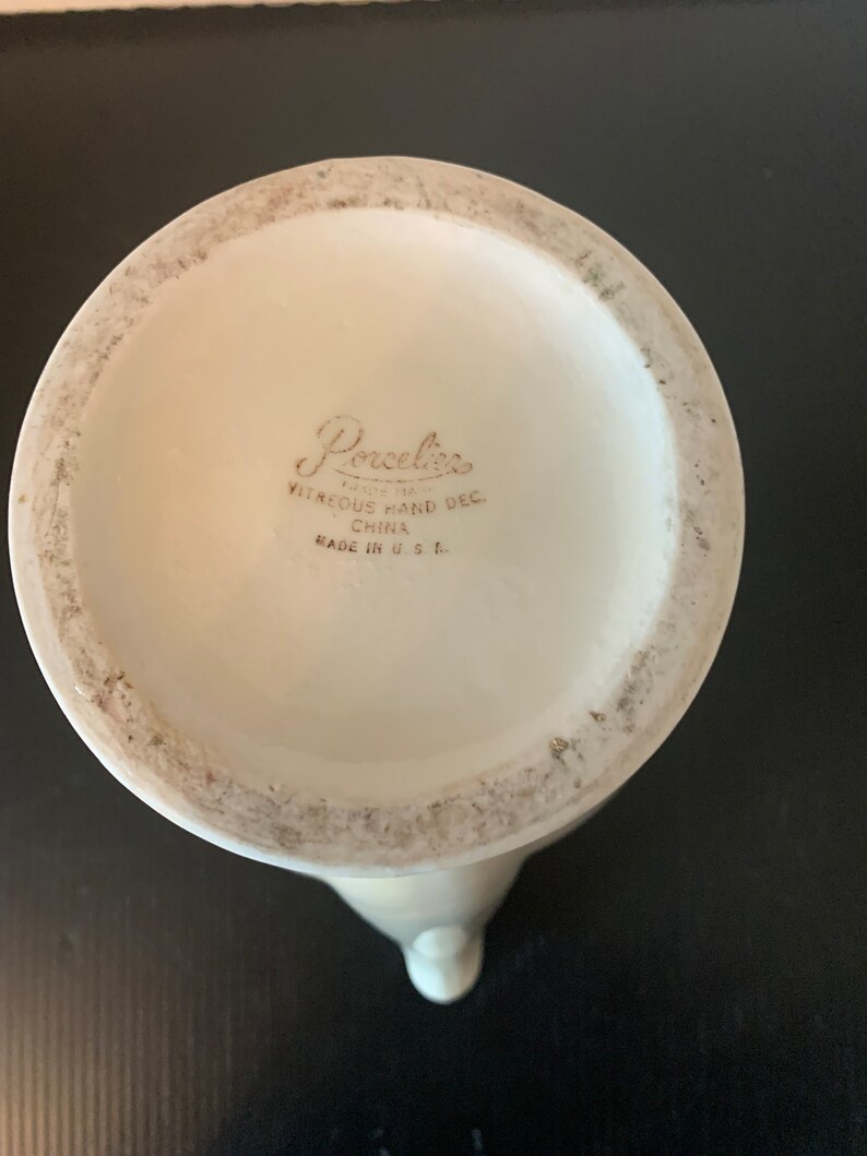 May include: The bottom of a white ceramic pitcher with the words "Porcelaine Since 1921 Vitreous Hand Dec. China Made in U.S.A." printed on the bottom.