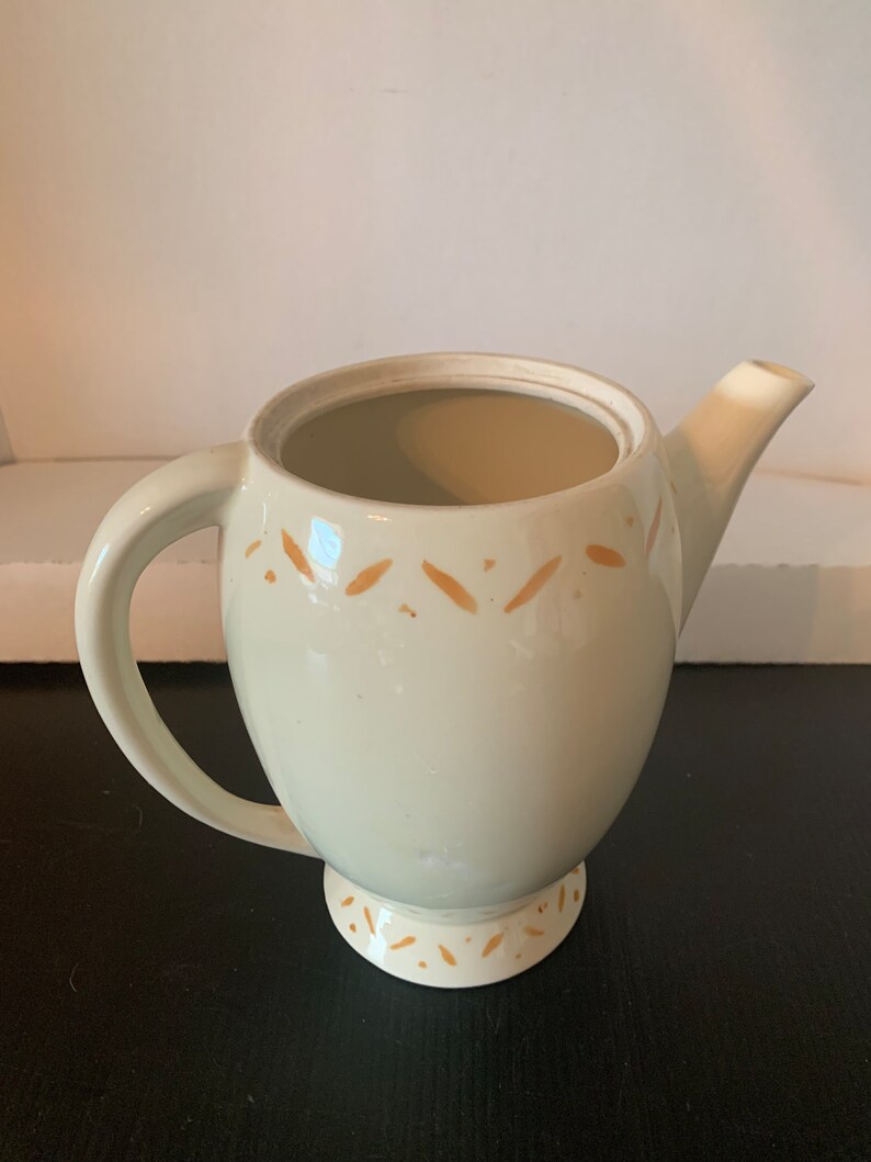 May include: A white ceramic teapot with a brown design. The teapot has a round body and a spout on the side. The teapot has a handle and a lid.