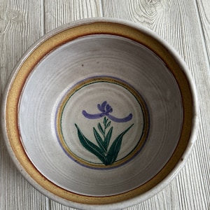 Puede incluir: Un tazón de cerámica blanca con un borde marrón y un diseño floral pintado a mano en tonos de morado, verde y amarillo.