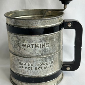 May include: A vintage silver metal sifter with a black handle and a black band around the top and bottom. The sifter has a crank on the top and says "WATKINS BAKING SPICES POWDER EXTRACTS" on the side.