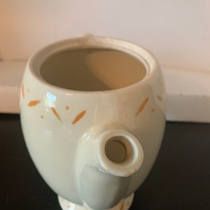 May include: A white ceramic teapot with a brown speckled design. The teapot has a round body and a spout. The teapot is empty.