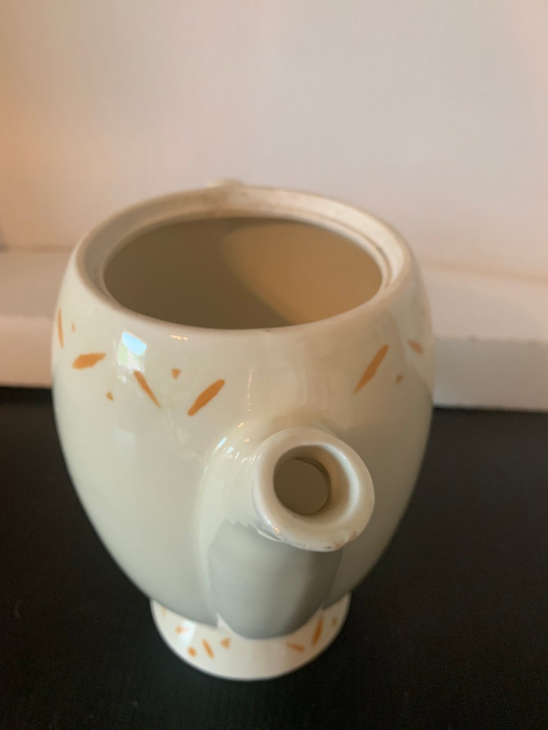 May include: A white ceramic teapot with a brown speckled design. The teapot has a round body and a spout. The teapot is empty.