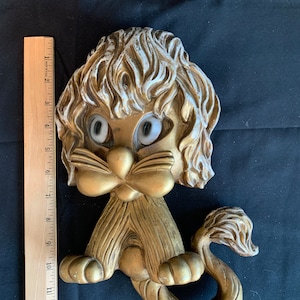 Puede incluir: Adorno de pared vintage con forma de león dorado con detalles blancos. El león tiene ojos grandes y de dibujos animados y una melena rizada. La pieza mide unos 33 cm de largo.