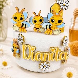 May include: A white cake decorated with yellow and blue bee-themed toppers, daisies, and the name "Dianita" in yellow. The cake is adorned with three bee figurines, a number one, and several daisy accents.