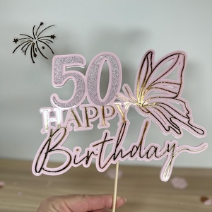 Könnte beinhalten: Ein rosa und goldener Glitzer-Kuchentopper mit der Zahl 50, den Worten "Happy Birthday" und einem Schmetterlingsdesign.
