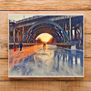 May include: A framed print of a winter scene featuring a bridge with arched supports over a frozen waterway. Silhouettes of people ice skating are visible. The sky is a warm orange and yellow, suggesting sunrise or sunset.