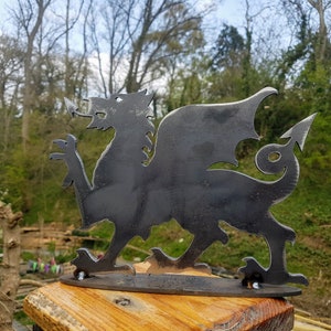 May include: A black metal silhouette of a dragon, the Welsh national symbol, mounted on a wooden base. The dragon is facing right with its wings spread out.