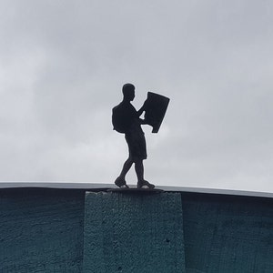 May include: Silhouette of a person with a backpack holding a map, standing on a wooden fence against a cloudy sky.