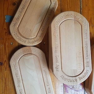 May include: Three wooden cheese boards with engraved text. The boards are oval shaped and have a raised edge. The text on the boards includes the names of different cheeses, such as Brie Camembert, Livarot, and Roquefort. The boards are made of light brown wood and have a natural finish.
