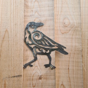 May include: A metal crow silhouette with a decorative, intricate design. The crow is attached to a wooden surface with a screw.