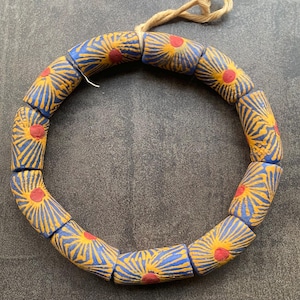 Puede incluir: Un collar de cuentas estampadas azules, amarillas y rojas. Cada cuenta cilíndrica tiene una base azul, un diseño de rayos de sol amarillos y un centro rojo. Las cuentas están ensartadas con un cordón de fibra natural.