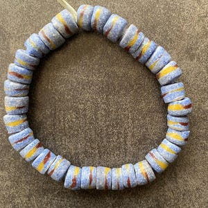 May include: A strand of blue, cylindrical beads with yellow and red stripes. The beads are strung together, forming a circular shape. The beads have a slightly textured appearance and are likely made of glass or a similar material.