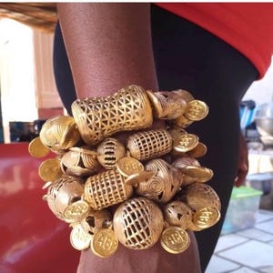 May include: A gold-toned bracelet featuring multiple cylindrical and spherical beads with intricate lattice and spiral patterns. The piece is accented with small, round, coin-like charms embossed with the letters ABP, creating a layered, textured look.